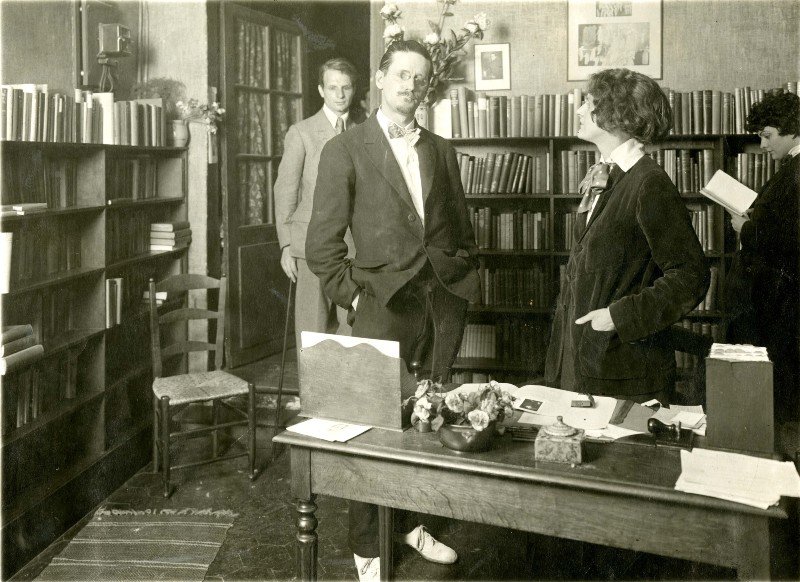 Old gray-scale image of a man standing behind a desk in a library with people browsing behind him